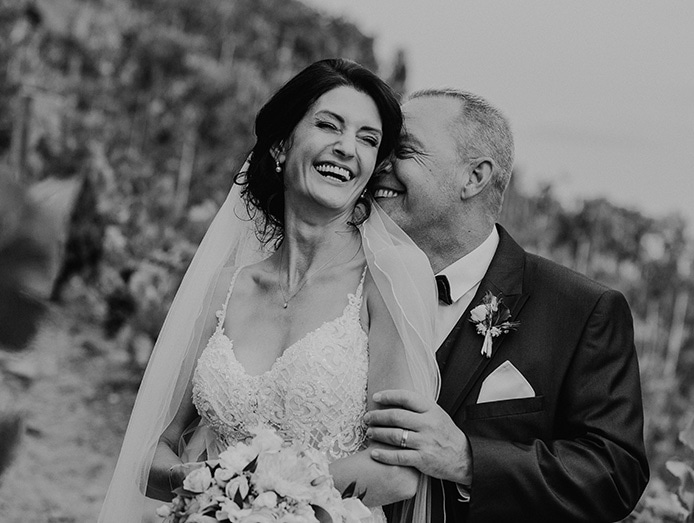 Schwarz-Weiß-Aufnahme eines lächelnden Brautpaares vor einem Weinberg