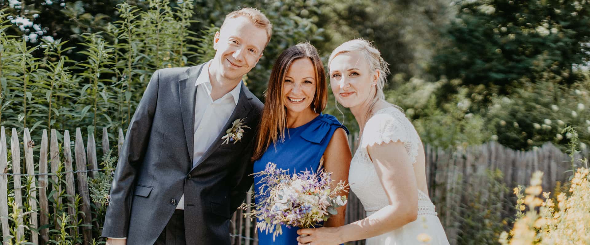 Brautpaar steht mit Kristin von festlicher vor einem Holzzaun im Sonnenschein und lächelt in die Kamera