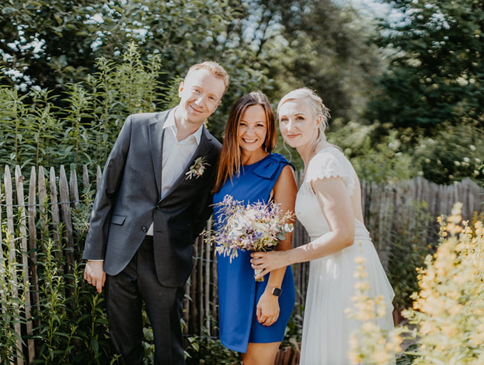Brautpaar steht mit Kristin von festlicher vor einem Holzzaun im Sonnenschein und lächelt in die Kamera