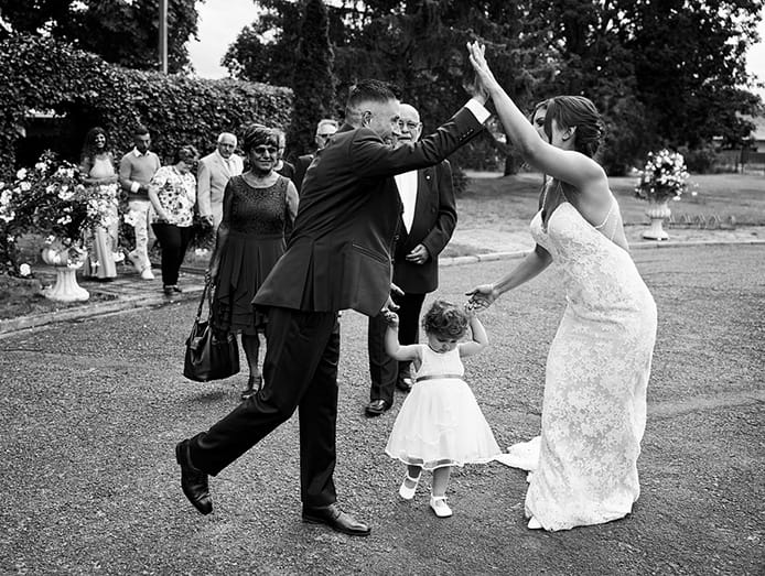 Schwarz-Weiß-Aufnahme eines Brautpaares, das sich ein High-Five gibt vor Hochzeitsgästen im Hintergrund