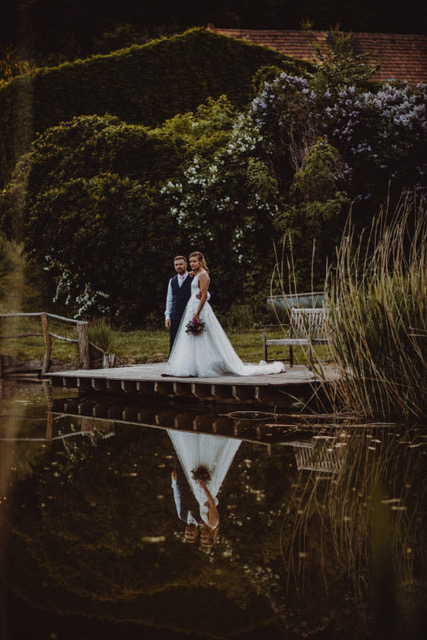 Brautpaar steht auf einem Holzsteg am Wasser und umarmt sich