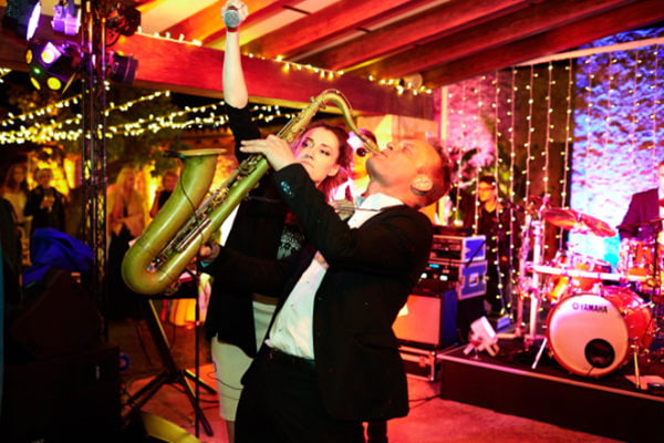 Frau steht mit Mikro neben spielendem Saxofonisten auf einer Bühne, Schlagzeug & Gäst im Hintergrund, bei beleuchteter Party-Atmosphäre