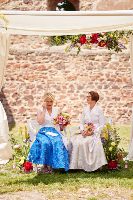 Gleichgeschlechtliches Brautpaar sitzt in der Sonne unter einem Sonnensegel mit jeweils einem Brautstrauß in der Hand auf Stühlen auf einer Wiese mit einer Klostermauer im Hintergrund