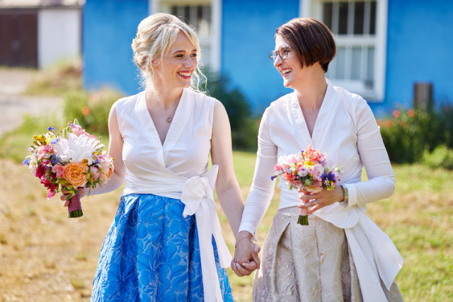 Gleichgeschlechtliches Brautpaar läuft über eine Wiese schaut sich fröhlich an, beide Bräute halten einen Brautstrauß in der Hand, Häuser im Hintergrund