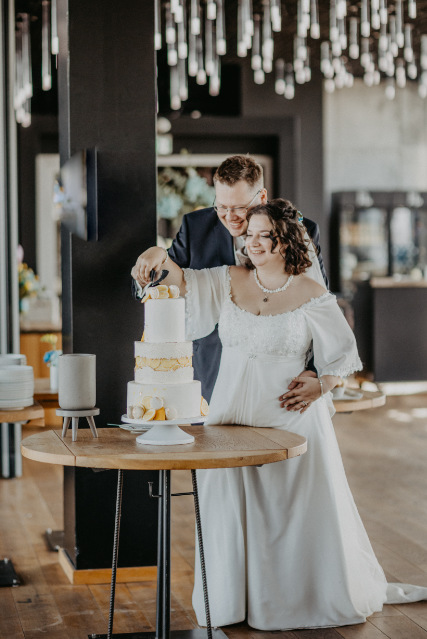 Brautpaar steht an einem kleinen runden Holztisch & schneidet gemeinsam die dreistöckige Hochzeitstorte an