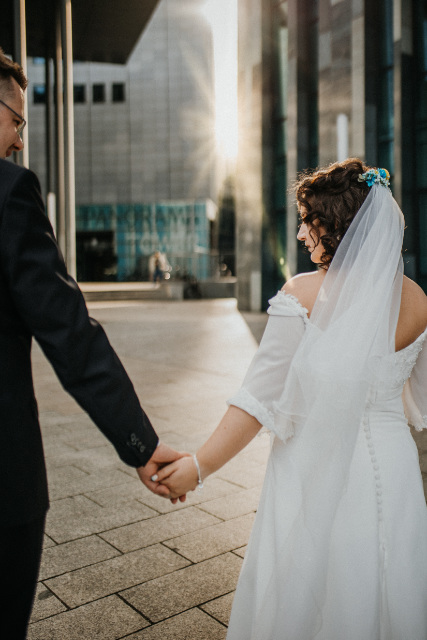 Brautpaar von hinten fotografiert Hand in Hand in auf Fußweg mit der Sonne im Hintergrund, die durch zwei Gebäude scheint
