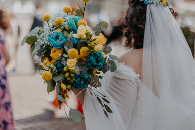 Braut von hinten fotografiert mit blau-gelbem Brautstrauß in der Hand