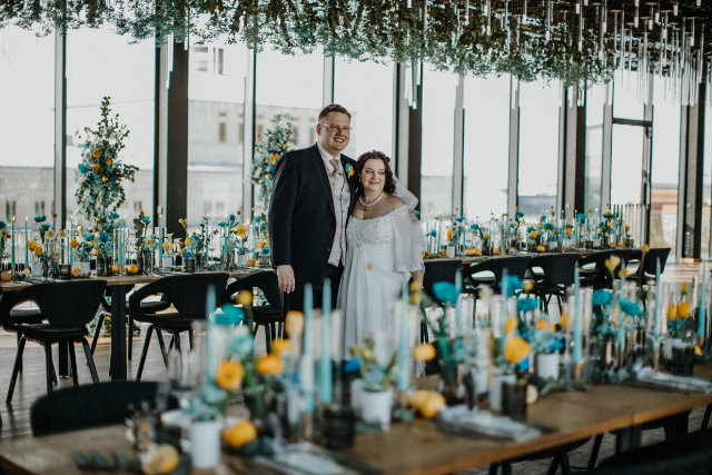 Brautpaar steht Arm in Arm & lächelnd zwischen blau-gelb dekorierten Tafeln mit großer Fensterfront im Hintergrund