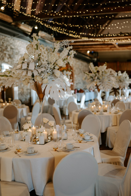 Weiß & dezent dekorierter Festsaalt mit Kerzen auf den Tischen, weißen Blumenstäußen & dezenter Lichterkettenbeleuchtung oben
