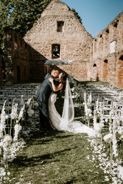 Brautpaar steht zwischen vielen weißen Stühlen auf der Wiese vor dem Altar & küsst sich, mit Klostergemäuer im Hintergrund