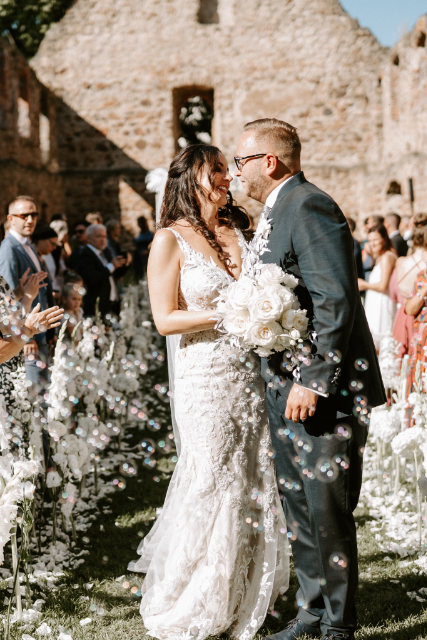Bautpaar steht in einem Seifenblasenregen & lächelt sich an während die Hochzeitsgäste Spalier stehen, Klostermauern im Hintergrund