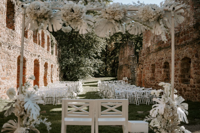Blick vom Altar zu den leeren Stuhlreihen der Hochzeitsgäste auf einer Wiese mit Bäumen im Hintergrund sowie links & rechts Klostermauern