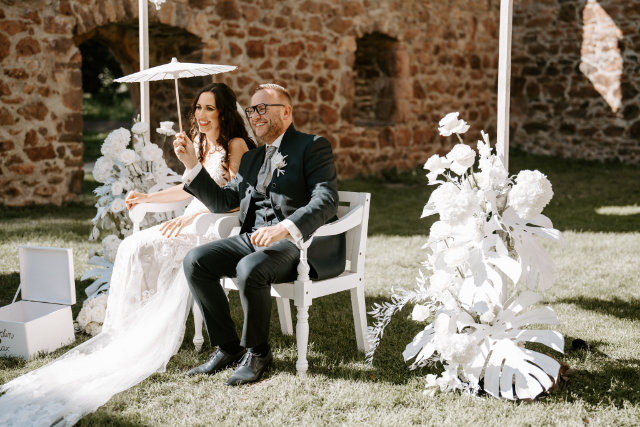 Brautpaar sitzt lächelnd auf zwei weißen Stühlen auf einer Wiese in der Sonne, Bräutigam hält der Braut einen kleinen Schirm, Klostermauern im Hintergrund